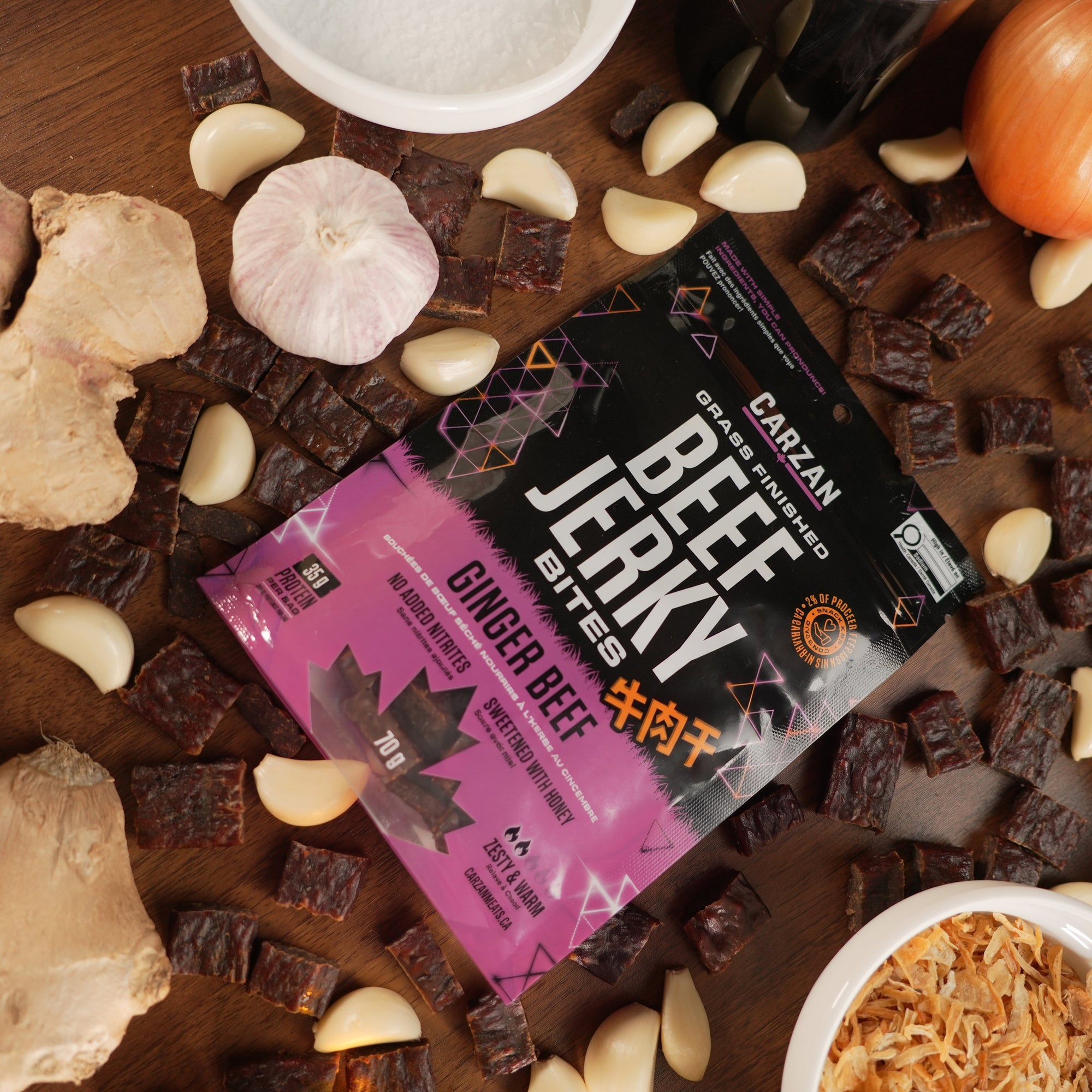 Carzan Ginger Beef grass-fed beef jerky bites bag surrounded by fresh ginger, garlic and onion, with loose jerky pieces on a wood table