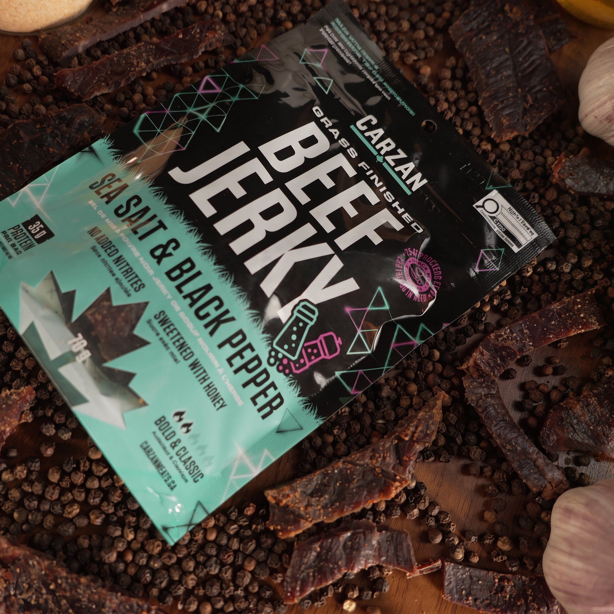 Carzan Sea Salt Black Pepper grass-fed beef jerky bag surrounded by peppercorns, garlic and onion, with loose jerky pieces on a wood table