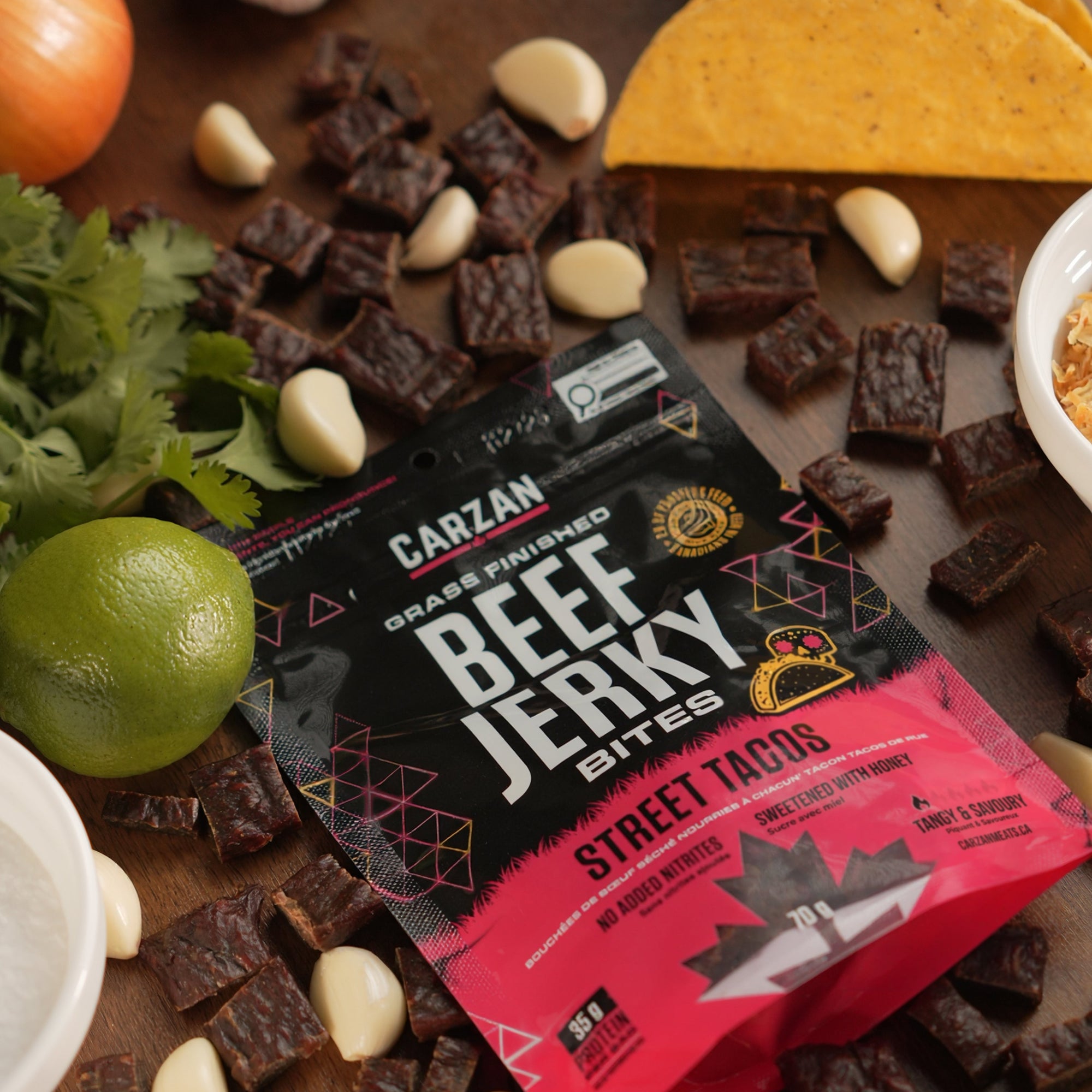 Carzan Street Taco grass-fed beef jerky bites bag surrounded by lime, cilantro, garlic and mini tacos, with loose jerky pieces on a wood table