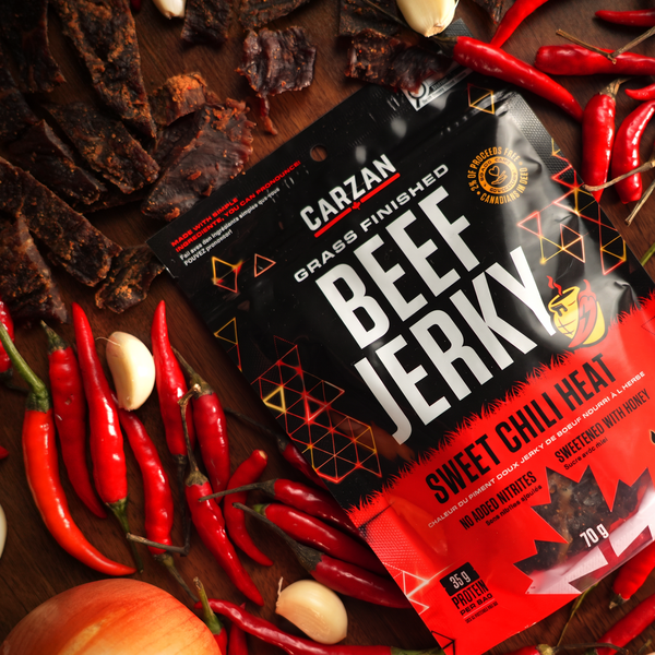 Carzan Sweet Chili Heat grass-fed beef jerky bag surrounded by fresh chilies, garlic and honey, with loose jerky pieces on a wood table