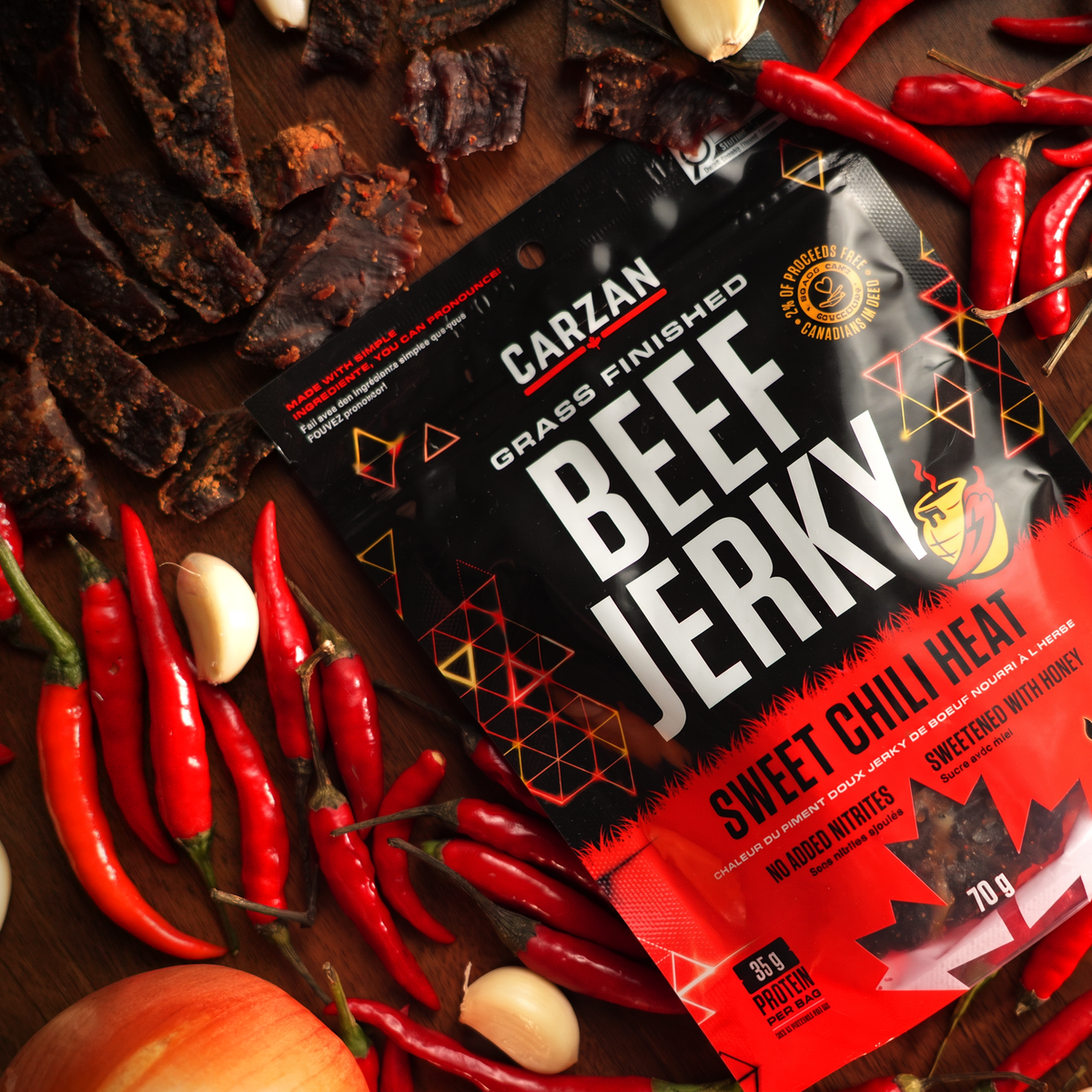 Carzan Sweet Chili Heat grass-fed beef jerky bag surrounded by fresh chilies, garlic and honey, with loose jerky pieces on a wood table